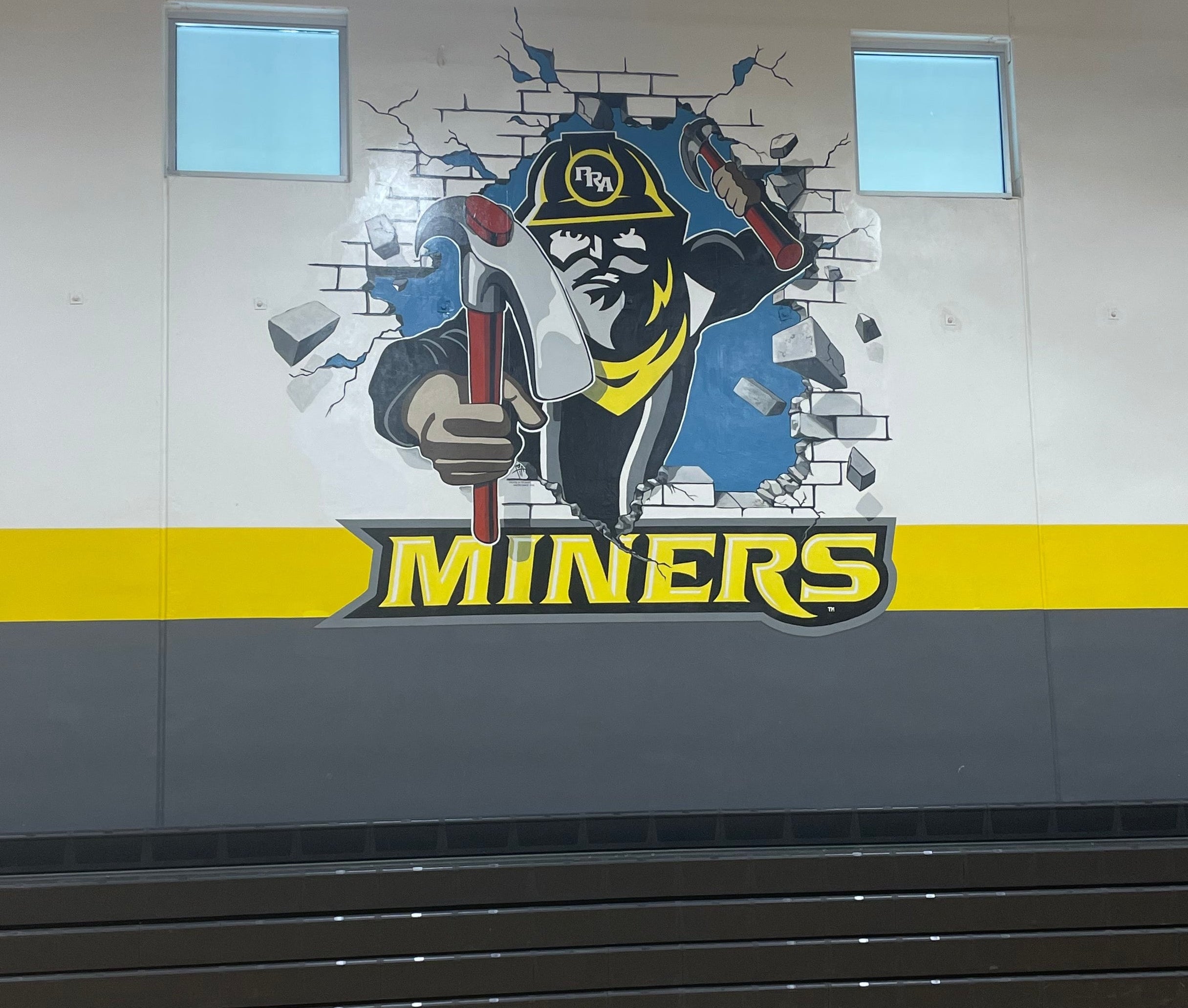 A large wall mural of a school mascot wearing a helmet with the word 'MINERS' below it, painted on an indoor gymnasium wall above a gray lower section and wooden court floor.